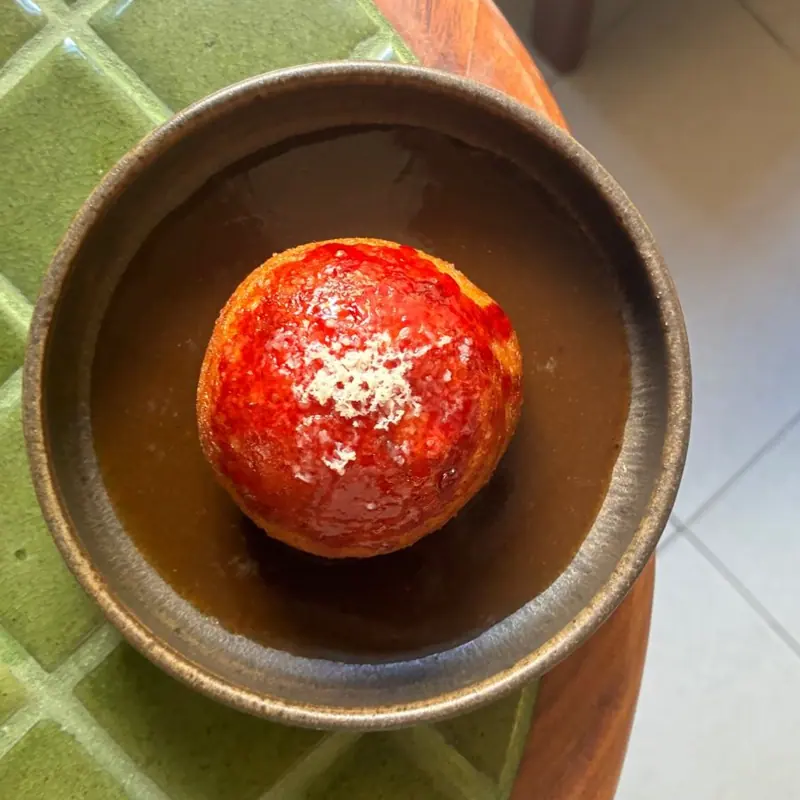 A fried ball topped with tomato sauce and grated cheese in a dark broth