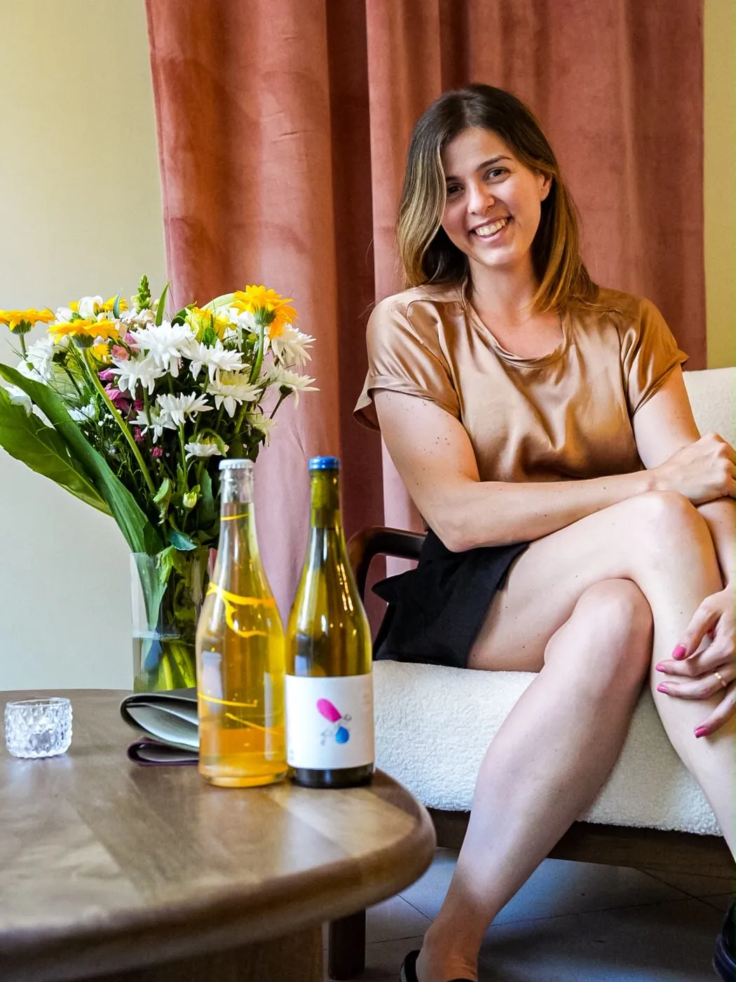 Vanessa Massé smiling in an armchair, beside two bottles of natural wine and a bunch of flowers